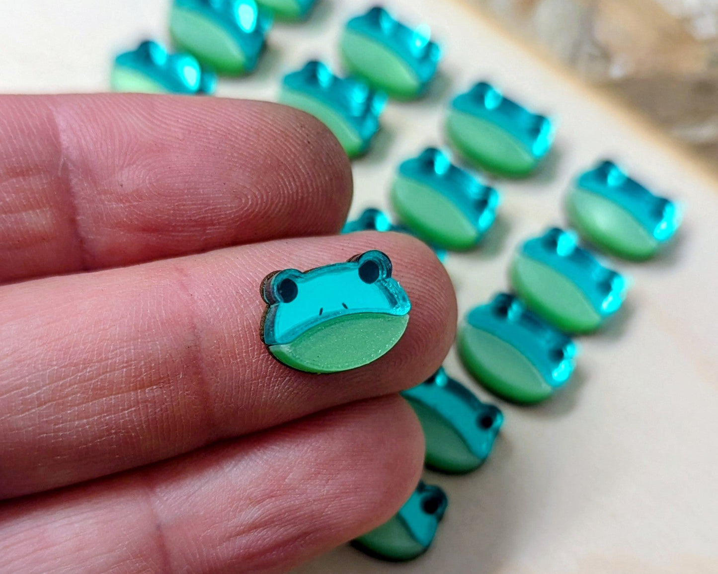 Small green frog-shaped earring held between fingers with a tray of similar objects in the background.