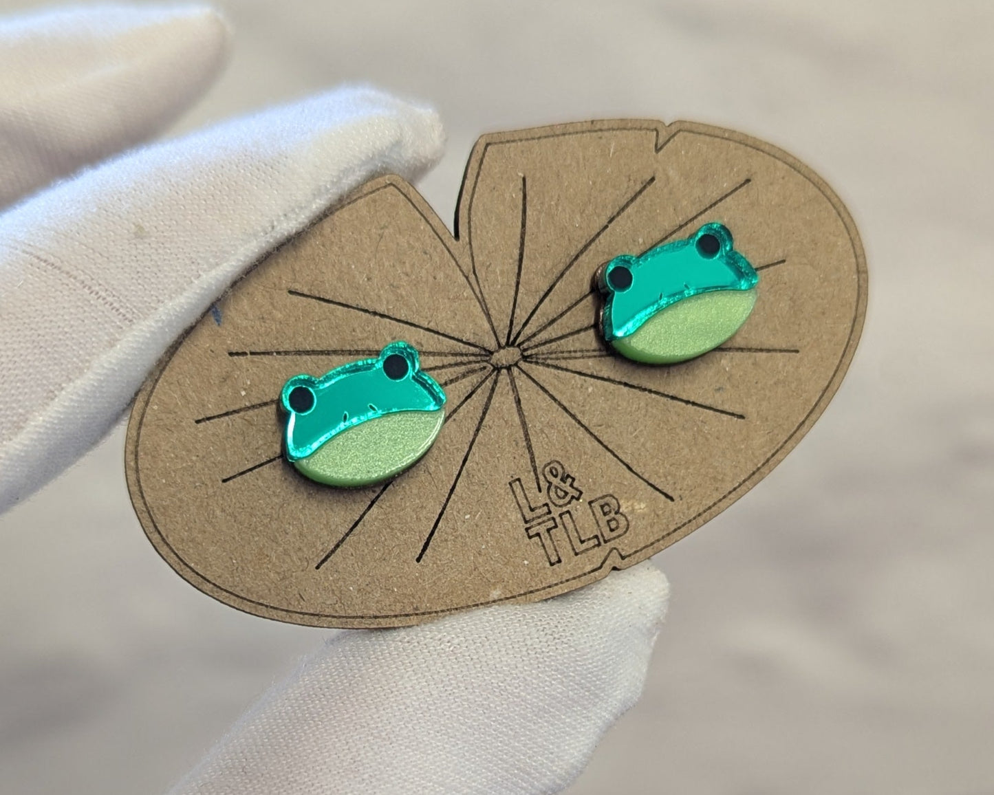 Pair of green frog-shaped earrings on a branded lily pad shaped card with 'L&TLB' written on.