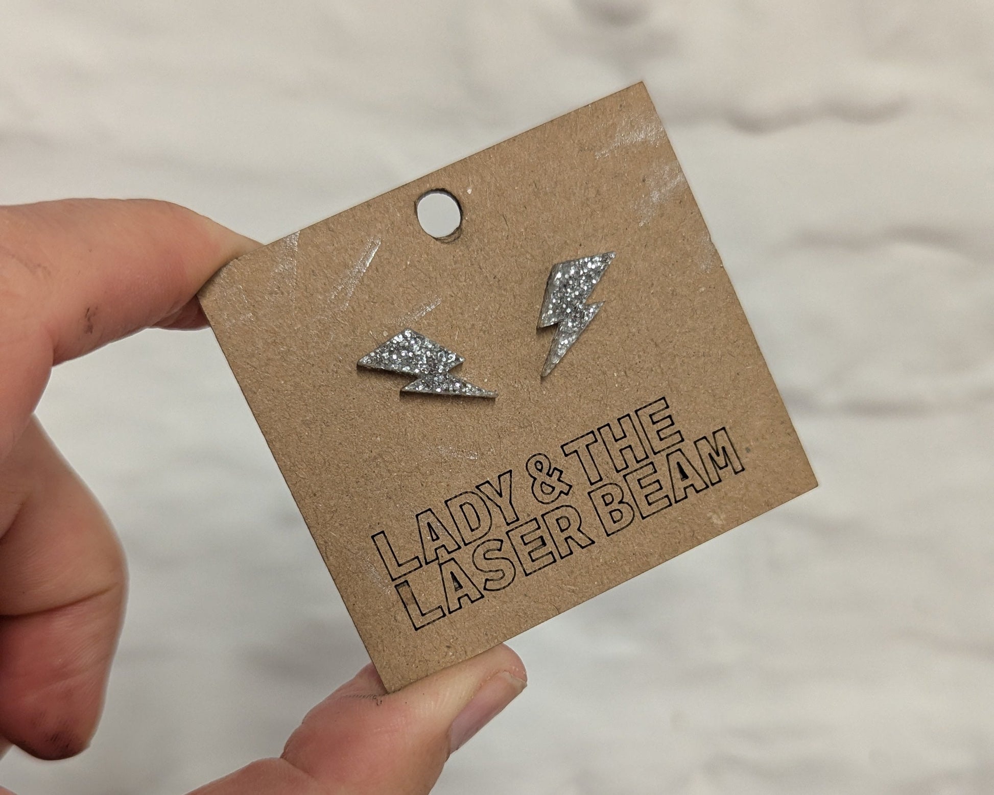 A hand holding a brown cardboard display card with two small silver lightning glitter stud earrings on it. The card has the text 'LADY & THE LASER BEAM' and is photographed against a light textured wall.