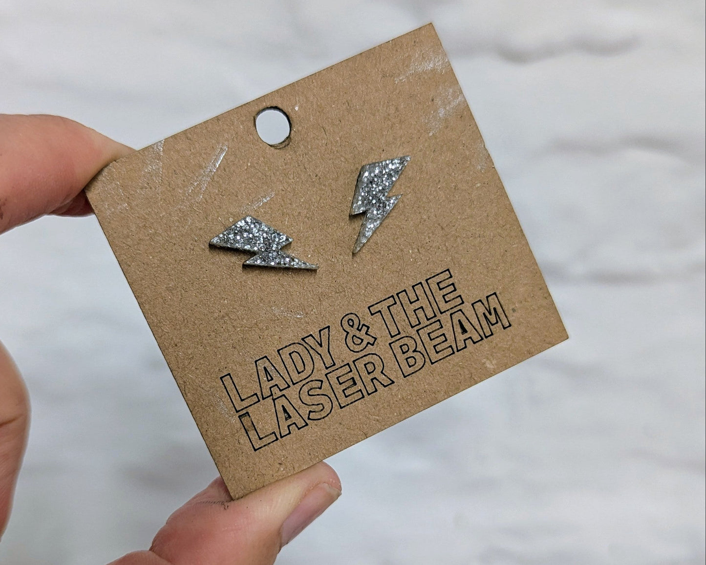 A hand holding a brown cardboard display card with two small silver glitterlightning bolt stud earrings. The card is printed with the brand name 'LADY & THE LASER BEAM'.