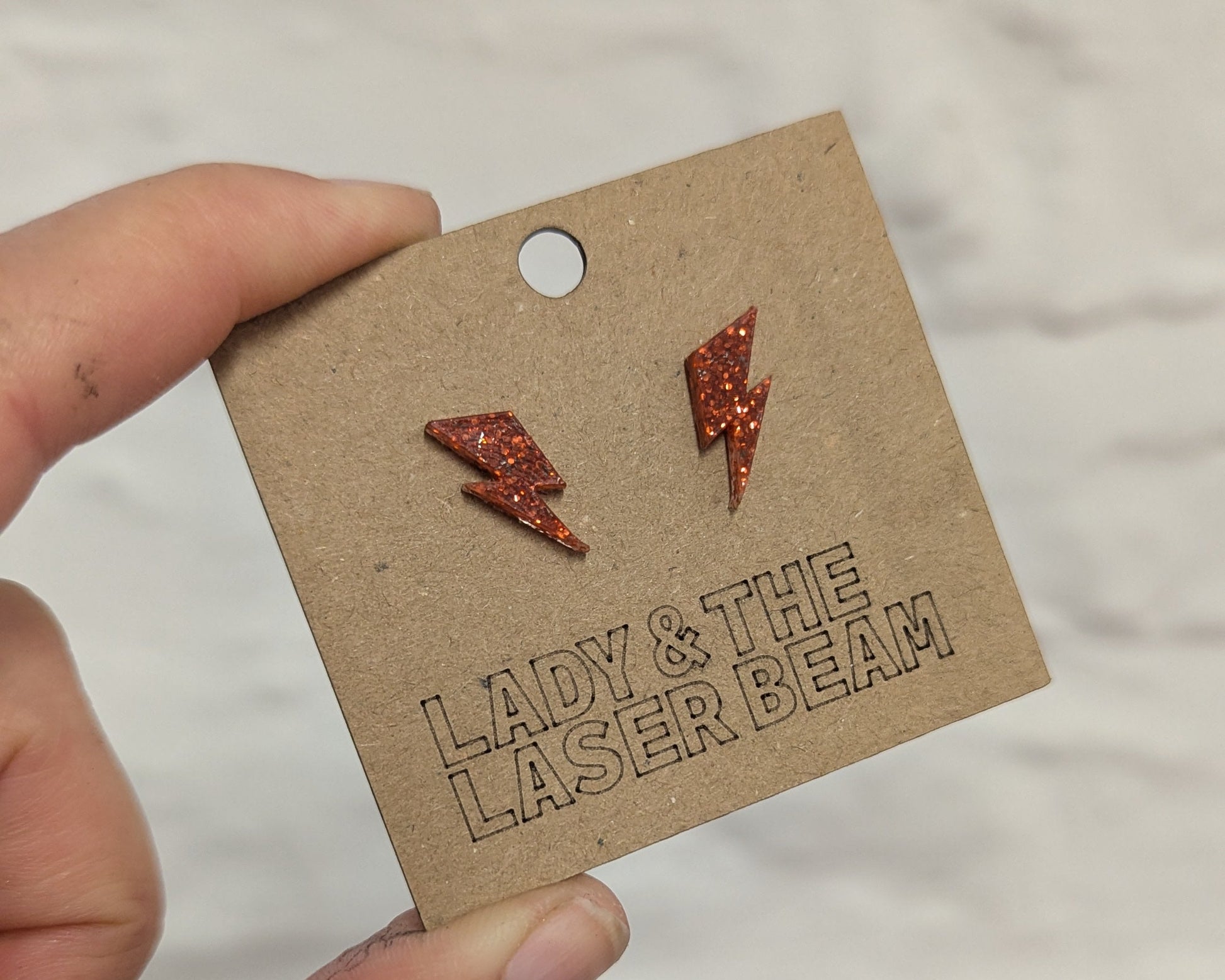 A hand holding a brown kraft display card featuring two small orange glitter lightning bolt stud earrings, with the brand name 'LADY & THE LASER BEAM' debossed on the card against a white textured background.