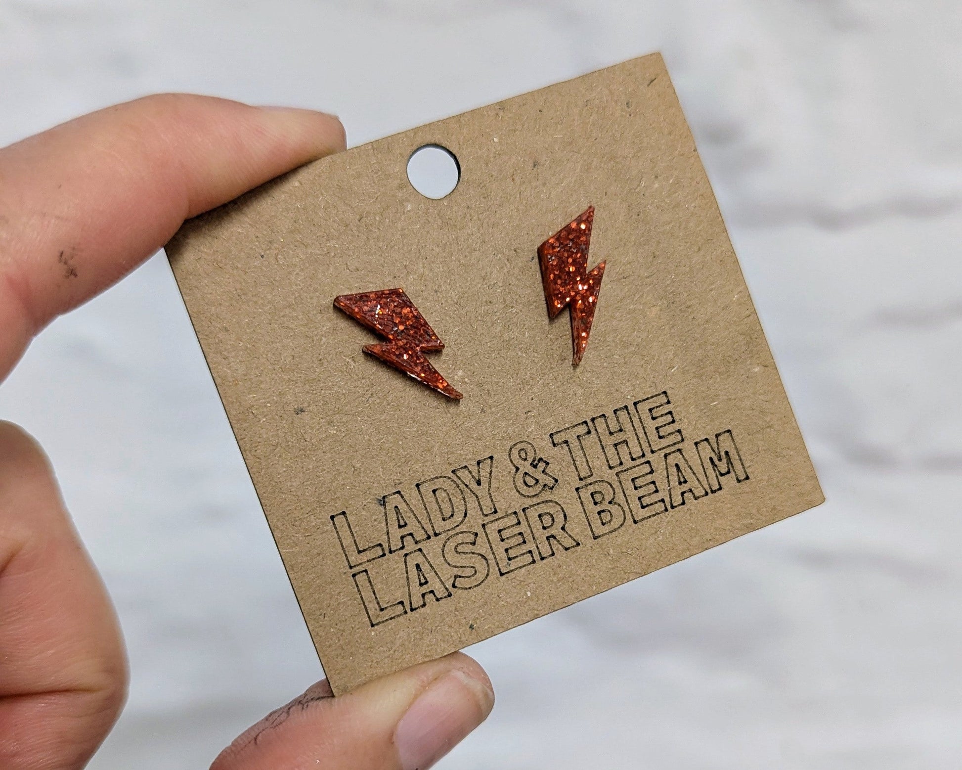A hand holding a brown kraft card displaying a pair of small orange glitter lightning bolt stud earrings, with the brand name 'LADY & THE LASER BEAM' printed on the card against a white textured background.