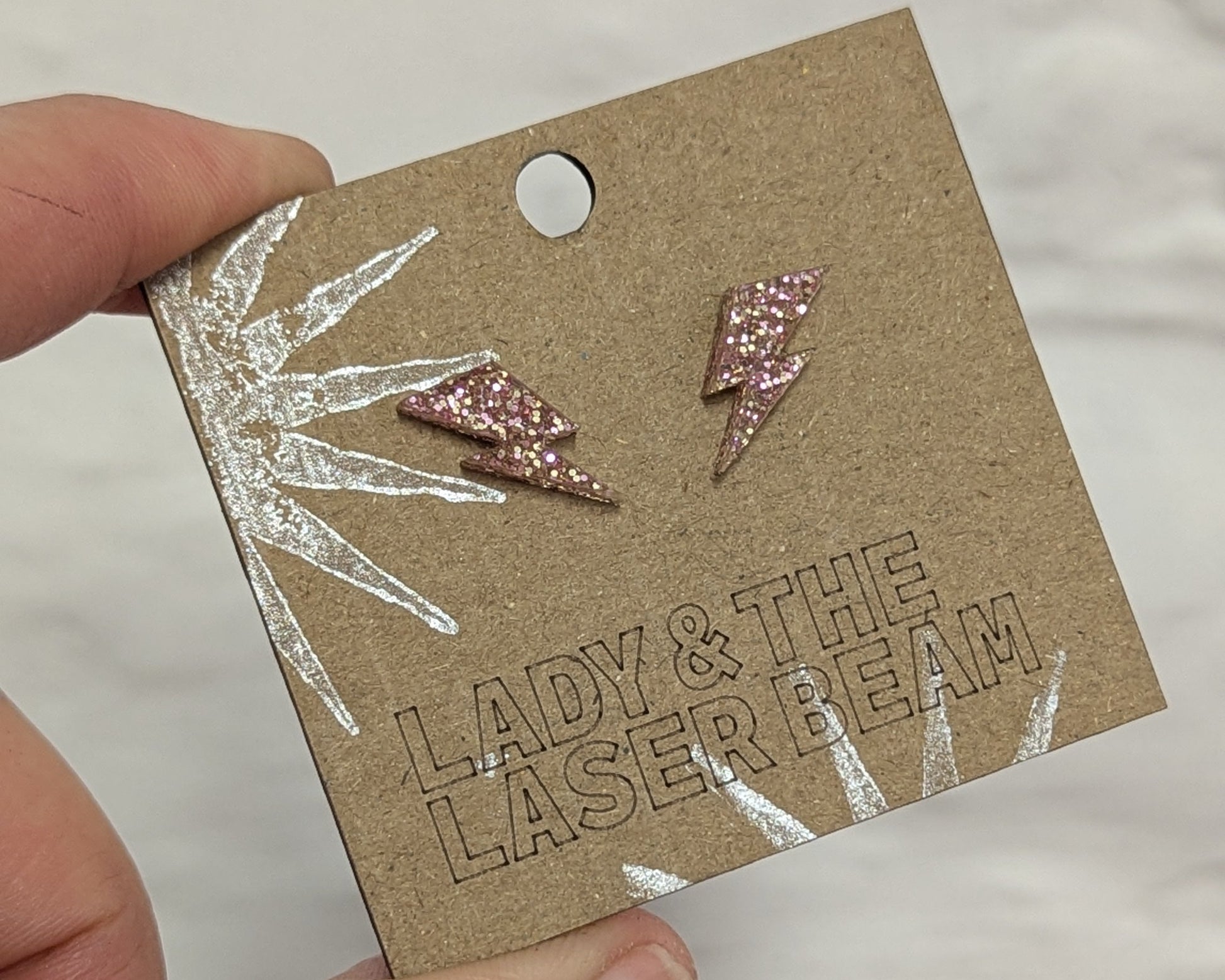 A hand holding a brown kraft jewelry display card with two small Rose Gold glitter lightning bolt stud earrings. The card has a cutout and the text 'LADY & THE LASER BEAM' debossed on it, with a silver foil leaf design on the left side.